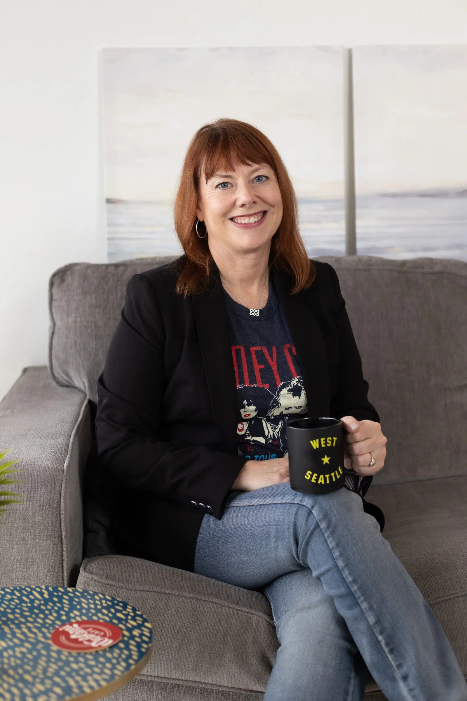 A woman with red hair sits on a gray couch, smiling, wearing a black blazer, jeans, and graphic T-shirt, holding a black mug that says "West Seattle," representing West Seattle web design creativity.