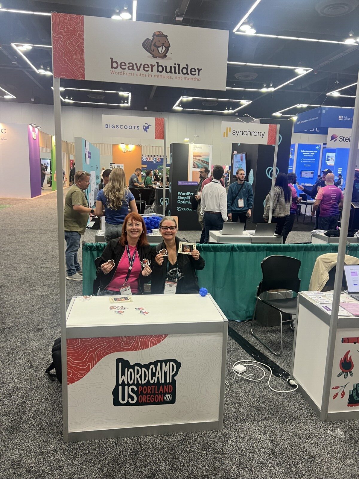 Two people smiling and holding items at a WordCamp event booth under a BeaverBuilder sign, showcasing the vibrant community at WordCamp US 2024.