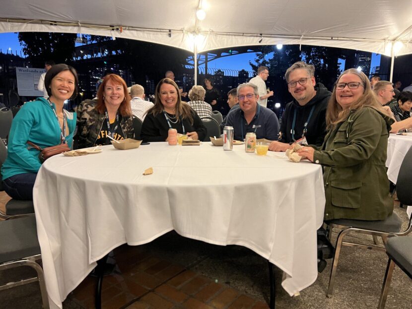 A group of six people sit around a round table under a white canopy at an outdoor event, smiling at the camera. They're enjoying their time at WordCamp US 2024, eager to share SEO 2024 insights and WordCamp lessons with fellow enthusiasts.