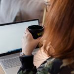 A person with red hair sits at a desk, holding a black mug while looking at a laptop screen, possibly exploring WordPress Web Design Services. A notebook and pen are also on the desk.