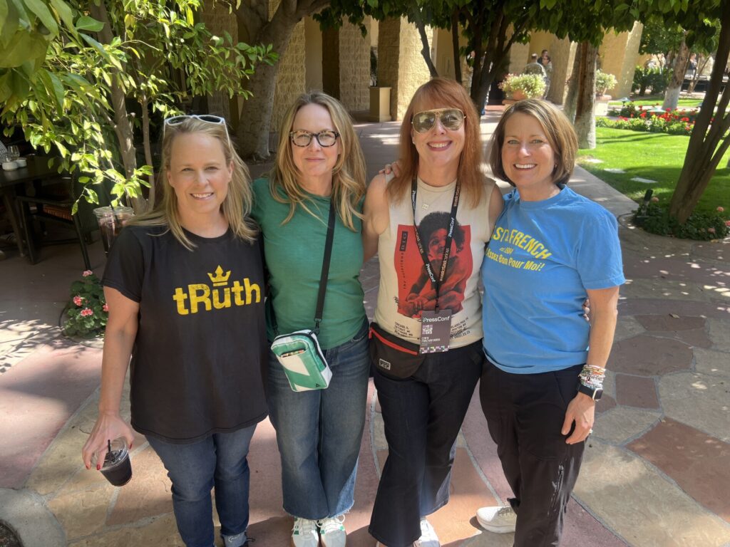 Four women stand outdoors on a stone path under trees, smiling at the camera for a year in review. Each wears casual clothes and sunglasses, with greenery and buildings visible in the background—a glimpse into 2025 moments together.