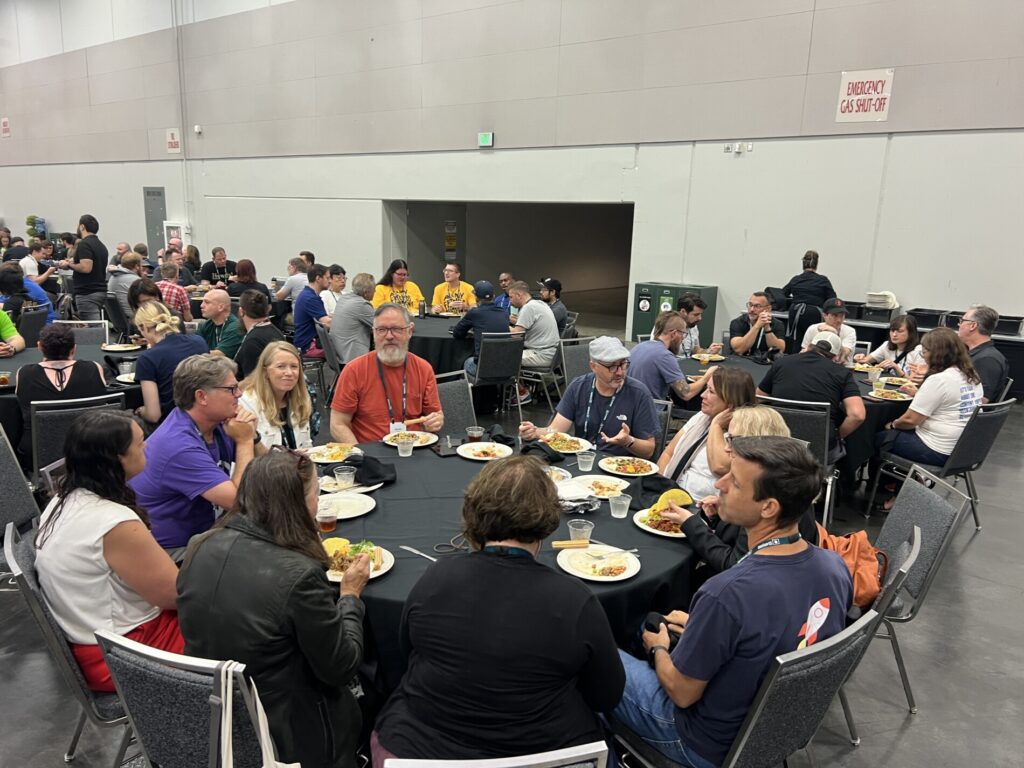 A large group of people are seated at round tables indoors, eating food and conversing during the 2025 year in review conference meal or event.