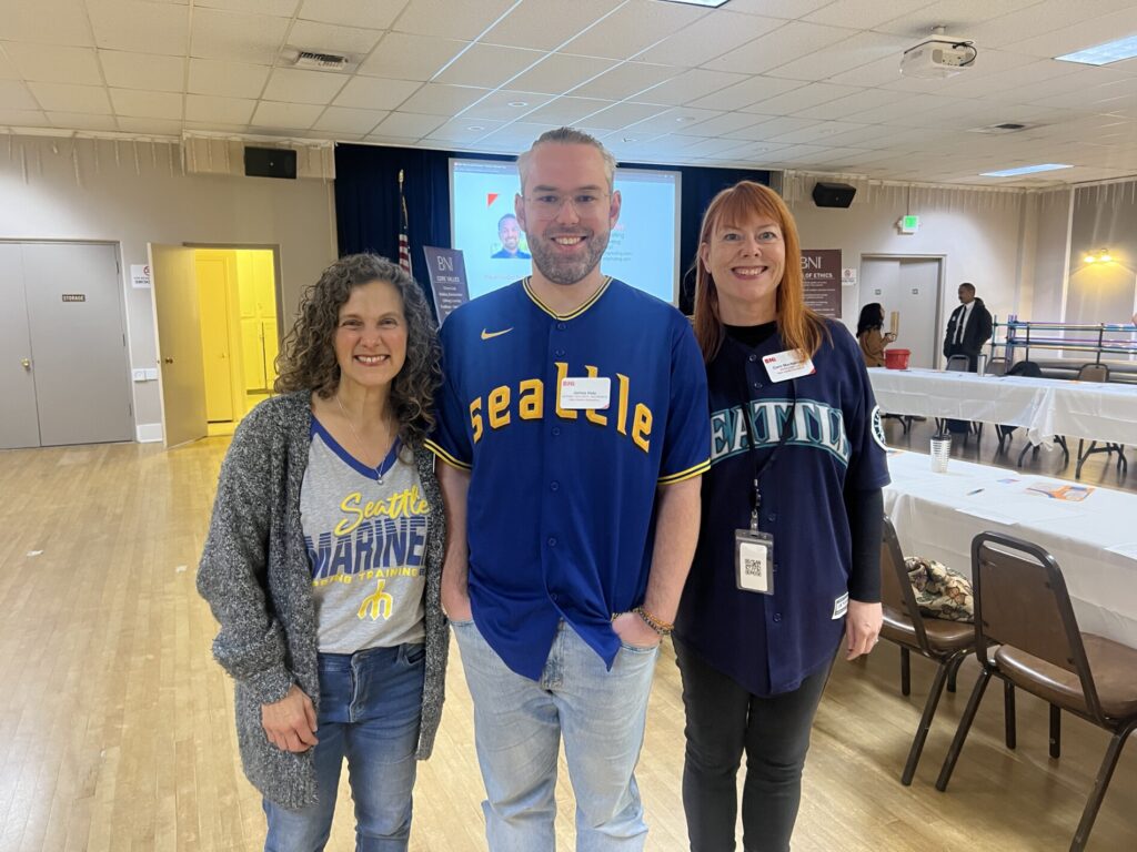 Three people stand indoors wearing Seattle Mariners shirts, smiling at the camera. Tables and chairs are visible in the background along with a projected screen displaying a 2025 year in review.