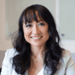 A West Seattle web designer with long dark hair and bangs is smiling, wearing a light blazer, against a blurred background.