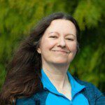 A woman with long brown hair and a blue jacket smiles confidently, standing outdoors against a green foliage background. Coach Denise Bodyworks radiates positivity and strength in her natural element.