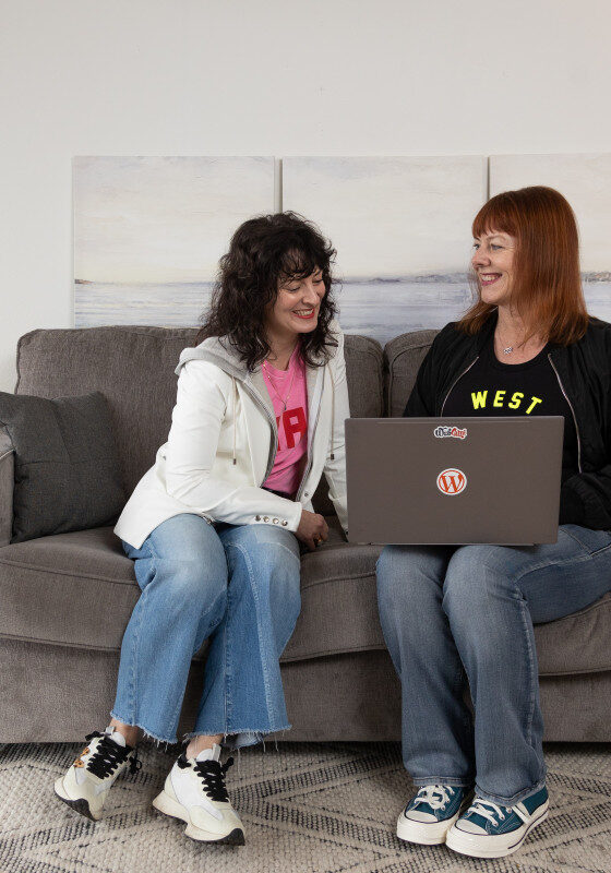 Two women sit on a gray couch, one holding a laptop while both smile—perhaps discussing WordPress Care Plans. A small table, plant, and floor lamp complete the cozy living room setting.