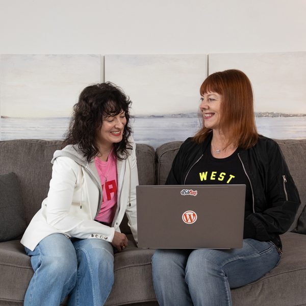 Two women sitting on a gray couch, smiling at each other while one works on a laptop adorned with stickers; the highly rated Seattle web designer is collaborating effortlessly.