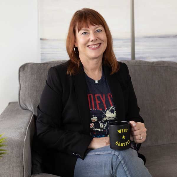 A woman with red hair sits on a grey couch, holding a black coffee mug labeled "West Seattle," wearing a black blazer and a graphic t-shirt that reads "Top Web Design Questions.