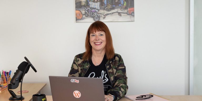 Cami MacNamara sits at a desk equipped with a laptop, microphone, and notebook. A colorful wall poster provides an eye-catching backdrop.