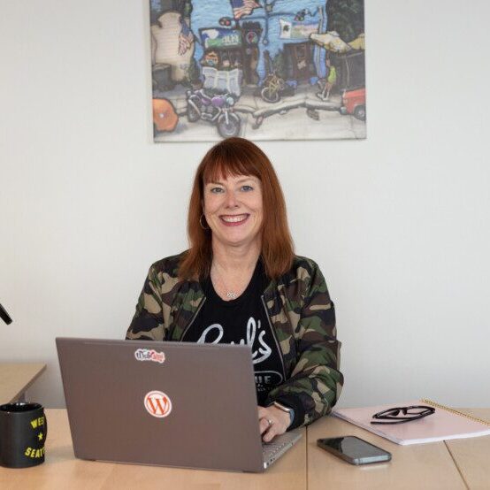 Cami MacNamara sits at a desk equipped with a laptop, microphone, and notebook. A colorful wall poster provides an eye-catching backdrop.