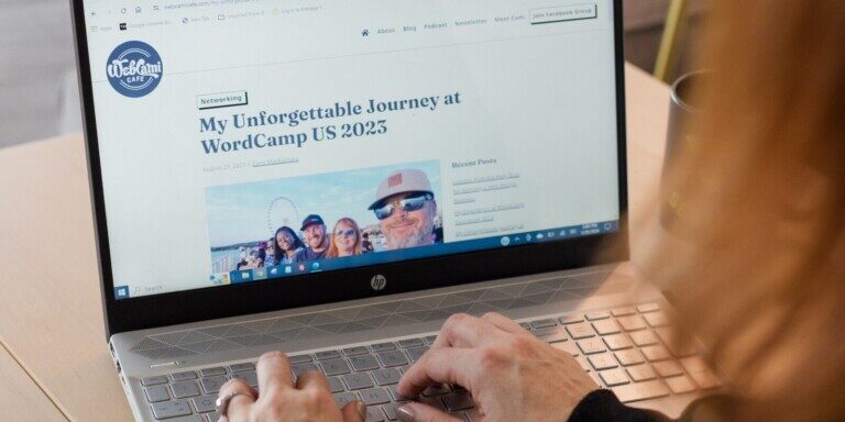 Cami MacNamara types on a laptop displaying a webpage titled "My Unforgettable Journey at WordCamp US 2023" with a notebook, pencil, and smartphone nearby, crafting tips for blogging success.