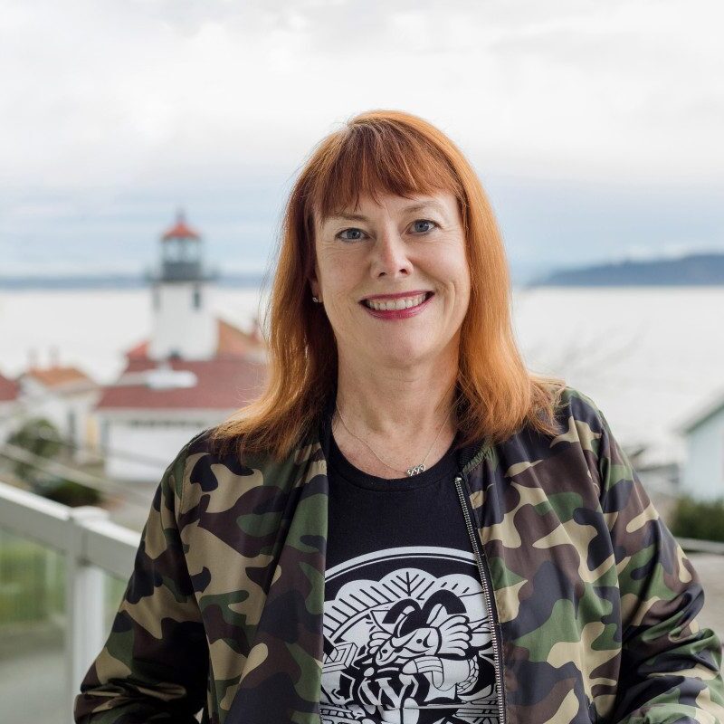 A woman with red hair wearing a camouflage jacket and black graphic t-shirt stands on a balcony, showcasing her Web Design Portfolio against a backdrop of houses and a lighthouse.
