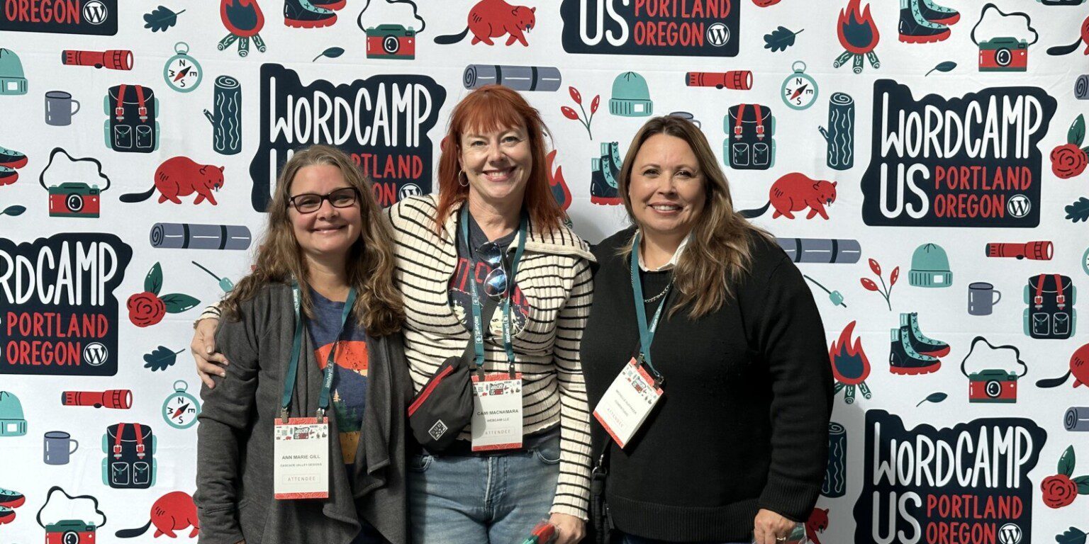 Three people standing together and smiling in front of a WordCamp US 2023 backdrop in Portland, Oregon. They are wearing nametags and holding small items, perhaps exchanging lessons for the upcoming 2024 event.