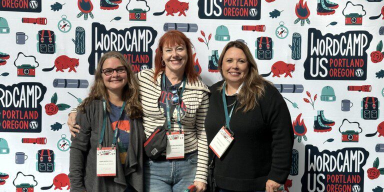 Three people standing together and smiling in front of a WordCamp US 2023 backdrop in Portland, Oregon. They are wearing nametags and holding small items, perhaps exchanging lessons for the upcoming 2024 event.