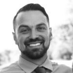 A West Seattle web designer with short dark hair and a beard, wearing a collared shirt and tie, smiles at the camera in an outdoor setting.