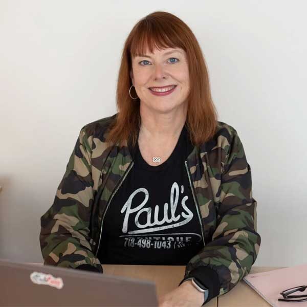 A woman with red hair wearing a camouflage jacket and a black "Paul's" t-shirt is sitting at a desk, showcasing her web design portfolio on a laptop while taking notes in a notepad.