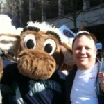 A person stands next to a mascot in a "Mariner" shirt, outdoors during the day, chatting about their work as a West Seattle web designer.