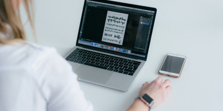         Person using a MacBook laptop with a calligraphy font design on the screen, adjusting the font size for optimal readability on their website. A smartphone rests on the table beside them, and they are wearing a smartwatch.