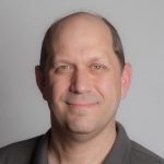 A man in a gray shirt is smiling for the camera in West Seattle.