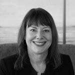 A woman with straight, shoulder-length hair smiles at the camera; she is wearing a dark blazer and sitting indoors, with a blurred background.